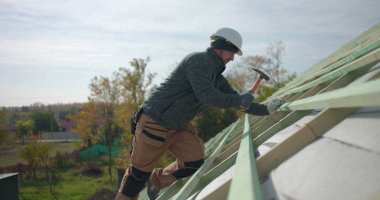 İnşaat işçisi inşaat yapısına çatı kirişleri çakıyor, güvenlik kaskı takıyor, arka planda iş arkadaşlarıyla takım çalışmasının yüksek açılı görüntüsü, açık hava inşaat projesi sitesi