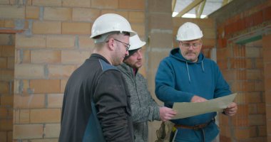 İnşaat işçileri inşaat planları, takım işbirliği, güvenlik miğferli üç adam tamamlanmamış tuğla duvar iç mimarisinin mimari planlarını inceliyor.