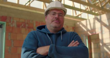 Güvenlik kaskı takmış, kollarını kavuşturan kendinden emin inşaat işçisi, inşaatta iskeleti, liderliği ve gücü olan tamamlanmamış inşaat sahasında duruyor.