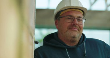 İnşaat işçisi kasketli, hafifçe gülümseyen, inşaat alanında duvara yaslanan, gevşek ifade, kapalı proje ayarları, doğal ışık