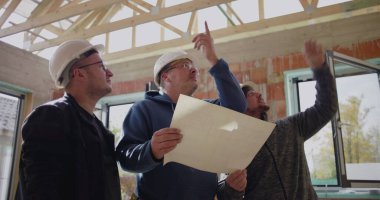 İyi aydınlatılmış bir inşaat alanının planlarını inceleyen inşaat işçileri, mimari planlara işaret ederek, proje detayları ve planlama üzerinde işbirliği yaparak,