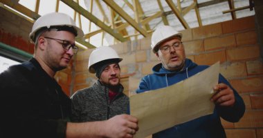 İnşaat işçileri kısmen inşa edilmiş yapının planlarını inceliyor, geniş bir inşaat alanında fikir alışverişinde bulunuyor ve proje detaylarını koordine ediyorlar.