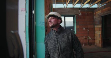 Dışarıya bakan genç inşaat işçilerinin yakın çekimi, sert şapkalı, odaklı ifade, kısmen inşa edilmiş tuğla duvarlı bir binanın penceresinin yanında duruyor.