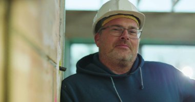 İnşaat işçisi kasketli, hafifçe gülümseyen, inşaat alanında duvara yaslanan, gevşek ifade, kapalı proje ayarları, doğal ışık