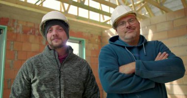 İnşaat halindeki bir binada yan yana duran iki inşaat işçisi, biri gülümsüyor diğeri ise kollarını kavuşturup takım çalışması ve çalışma arkadaşlığı yapıyor.