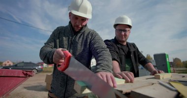 İki inşaatçı birlikte çalışıyor, biri odun kesmek için el testeresi kullanırken diğer saatler, etkin inşaat sahasında takım çalışması, yapı iskeleti ve aletler etrafında.