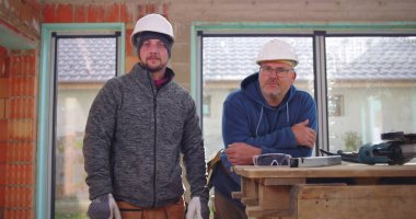 İnşaat işçileri inşaat halindeki çalışma tezgahının yanında gülümsüyor, miğferler takılı, takım çalışması ve inşaat sahasında dostluk, pozitif işyeri atmosferi