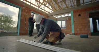 İnşaat işçisi, binanın iç kısımlarındaki PVC borularını diz çöküp ölçerken, arka plandaki meslektaşları da projeleri, inşaat alanındaki takım çalışmalarını gözden geçiriyorlar.