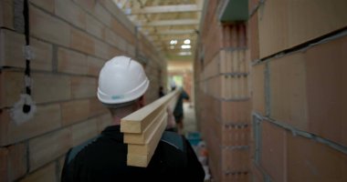 İnşaat işçisi omuzunda tahta kalaslar taşıyor, bitmemiş bir binada dar koridorlarda yürüyor, inşaat alanında takım çalışması ve işçilik yapıyor.