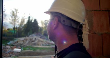 İnşaat işçisi kasketli, gülümseyen, dışarıdan bakan, yan profilli, doğal çevresi olan, inşaat alanında rahat tavırlı biri.