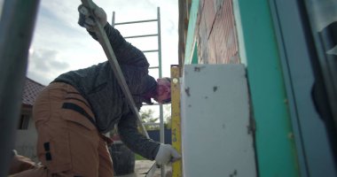 İzolasyon tesisatı üzerinde çalışan, iskele üzerinde diz çöken, kesinlik için seviye aletini kullanan, inşaat halindeki konut evinin dış duvarına odaklanmış bir yapı.