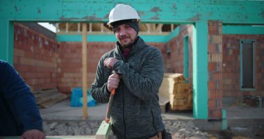 İnşaat işçisi elinde çekiçle inşaat alanında güvenle gülümsüyor takım çalışması ve inşaat halindeki konut yapısına olan bağlılığı profesyonel inşaatçı.