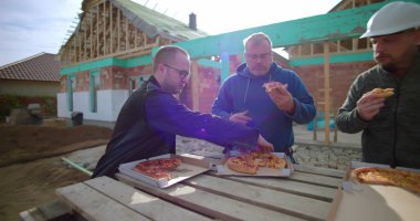 İnşaat ekibi öğle arasında pizza yiyor, derme çatma masa etrafında toplanıyor, arka planda tamamlanmamış bina yapısı, takım çalışması ve günlük açık hava yemeği.