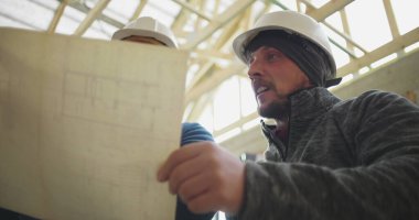 Kapalı alandaki mimari planı gözden geçiren inşaat işçisi, iş arkadaşlarıyla bina planlarını tartışıyor, inşaat projesinde takım çalışması sürüyor