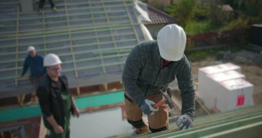 Meslektaşlar gözlemlerken inşaat işçisi çatı kirişlerini çekiçlerken, yüksek yapı üzerindeki takım çalışması, açık hava projesine odaklanmış eylemlerin yakın plan çalışması