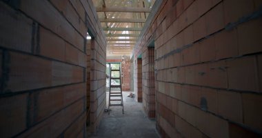 Açığa çıkmış tuğla duvarları, ahşap tavan kirişleri ve kapı eşiğine yerleştirilmiş bir merdiveni olan tamamlanmamış binanın iç manzarası.