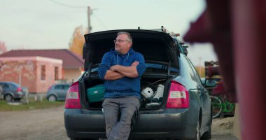 İnşaat alanındaki araba bagajına dayanan işçiler, kollar çapraz ve çevreyi gözlüyor, yenileme projesi sırasında düşünceli ve hazırlıklı olunduğunu vurguluyor