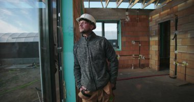 Dışarıya bakan genç bir inşaat işçisi, elleri ceplerinde, kaskı takılı, kısmen inşa edilmiş binanın içinde, tamamlanmamış tuğla duvarlar ve arka planda pencere çerçevesi var.