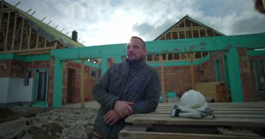 İnşaat alanındaki ahşap paletlere yaslanan adam, yanına yerleştirilmiş kask ve eldivenler, güneş ışığıyla uzaklığa bakıyor, arka planda kısmen inşa edilmiş bir ev.