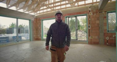 İnşaat işçisi miğferiyle inşaat halindeki evde yürüyor, ilerlemeyi gözlemliyor, etrafı tuğla duvarlar, ahşap kirişler ve pencere çerçeveleriyle çevrili.