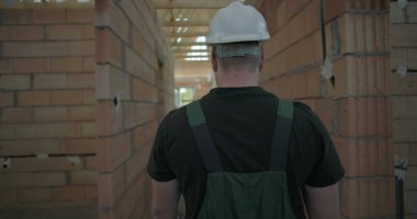Erkek işçi, tamamlanmamış binanın koridorunda yürüyor, miğfer takıyor, etrafı inceliyor, tuğla duvarlar ve ahşap iskeletle kaplı.