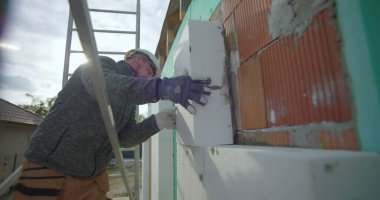 Dış duvara yalıtım paneli yerleştiriliyor, eldiven ve kask takan inşaat işçisi tuğla cephesine baskı yapıyor, konut yenilemesi yapıyor.