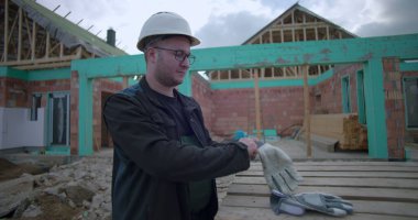 İnşaat alanında eldiven takan, güvenlik kaskı ve gözlük takan, arka planda kısmen inşa edilmiş evi olan ahşap paletlerin yanında duran bir adam.