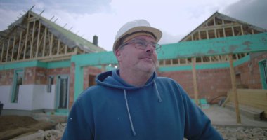 Adam şantiyeye dikkatlice bakıyor, güvenlik kaskı ve gözlük takıyor, arka planda kısmen inşa edilmiş bir ev var.