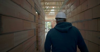 Tamamlanmamış binanın koridorunda yürüyen, güvenlik kaskı ve gözlük takan, tuğladan duvarları ve ahşap iskeleti olan bir erkek işçi.