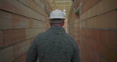 İnşaat işçisi miğferiyle bina içini teftiş ediyor, dikiz aynasından bakıyor, ahşap merdiveni ve tamamlanmamış tuğla duvarları aşıyor.