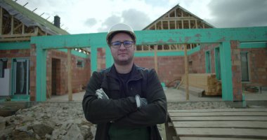 İnşaat alanında kolları çapraz duran adam, güvenlik kaskı ve gözlük takıyor, kısmen inşa edilmiş ev ve arka planda ahşap paletler var.