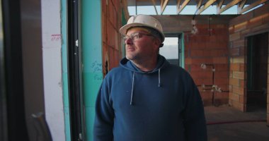 Olgun bir inşaat işçisi, düşünceli bir şekilde bakıyor, kısmen inşa edilmiş binanın içinde, tavan kirişleri ve tuğla duvarları açık.