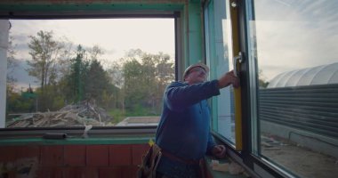 İşçi inşaat alanında pencere çerçevesinin hizalanmasını sağlamak için seviye aracı kullanıyor, sabit kasklı odaklı işçinin yakın plan pencere açılışı yakınlarındaki ölçümleri ayarlıyor