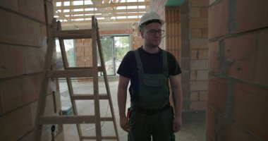 Erkek işçi bitmemiş binanın koridorunda yürüyor, miğfer ve gözlük takıyor, tuğla duvarları ve ahşap tavan iskeletini inceliyor.