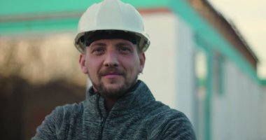Başlık takan inşaat işçisi, inşaat alanında güvenle duran, kolları çapraz duran, açık yapının arka planı, inşaattaki güvenilirliği, gururu ve uzmanlığı sembolize eden,