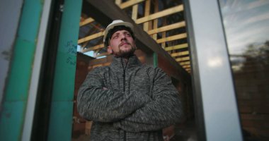 Kolları çapraz genç inşaat işçisi, sıkı bir şapka takıyor, kararlı görünüyor, bir inşaat alanının arka planında ahşap iskeleti olan cam bir duvarın yanında duruyor.