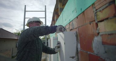 Dış duvardaki yalıtım paneli, güvenlik miğferi olan inşaat işçisi tuğla cephesine baskı yapıyor, bina yalıtımı...