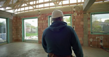 Tamamlanmamış binanın koridorunda yürüyen, güvenlik kaskı ve gözlük takan, tuğladan duvarları ve ahşap iskeleti olan bir erkek işçi.