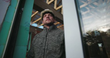 Sert şapkalı, yansıtıcı ifadeli, cam ve ahşap iskeletin yanında duran genç inşaat işçisi, proje inşaatına odaklandı