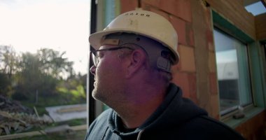 Sert şapkalı, açık havada duran, kapının yanında duran, arka planda doğal çevresi ve enkazı olan olgun inşaat işçilerinin yan profili.