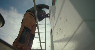 Dış duvara yalıtım paneli yerleştiriliyor. İnşaat işçisi iskelede çalışıyor. Tam konum için elleri kullanıyor.