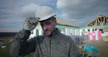 Başlık takmak, kameraya gülümseyen inşaat işçisi, arka planda inşaat alanı, profesyonel ve cana yakın görünüm.