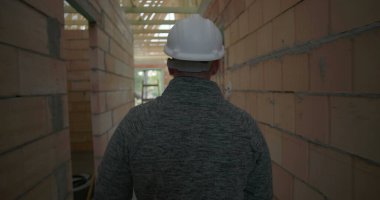 İnşaat işçisi miğferiyle bina içini teftiş ediyor, dikiz aynasından bakıyor, ahşap merdiveni ve tamamlanmamış tuğla duvarları aşıyor.