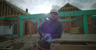 İnşaat alanında ahşap paletlere yaslanan, güvenlik kaskı ve eldiven takan, uzaktan bakan, kısmen inşa edilmiş arka planda evi olan bir adam.