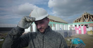 Başlık takmak, kameraya gülümseyen inşaat işçisi, arka planda inşaat alanı, profesyonel ve cana yakın görünüm.