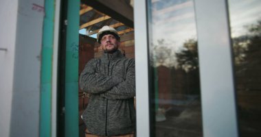 Kolları çapraz genç inşaat işçisi, sıkı bir şapka takıyor, kararlı görünüyor, bir inşaat alanının arka planında ahşap iskeleti olan cam bir duvarın yanında duruyor.