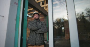 Kolları çapraz genç inşaat işçisi, sıkı bir şapka takıyor, kararlı görünüyor, bir inşaat alanının arka planında ahşap iskeleti olan cam bir duvarın yanında duruyor.