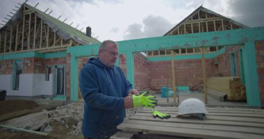 İnşaat alanında eldiven ve miğfer takan bir adam, arka planda kısmen inşa edilmiş bir evin yanında duruyor, dışarıda çalışmaya hazırlanıyor.