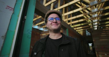 Sert şapka ve gözlüklerle gülümseyen inşaat işçisi, inşaat alanında açık çatı çerçevesi altında güvenle duruyor, pozitif tavır sergiliyor ve projeye odaklanıyor