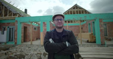 İnşaat alanında kolları çapraz duran adam, güvenlik kaskı ve gözlük takıyor, kısmen inşa edilmiş ev ve arka planda ahşap paletler var.